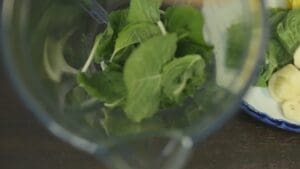 Fresh spinach leaves in a blender with sliced banana and pineapple on a plate nearby