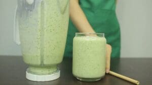 A person places a glass of green smoothie next to a blender filled with the same mixture
